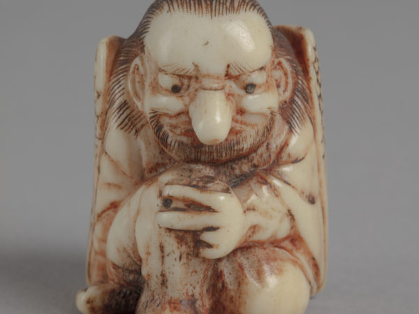 A Japanese ivory netsuke of a seated tengu. It has the body of a man, the wings of a bird, a beard and a long nose.