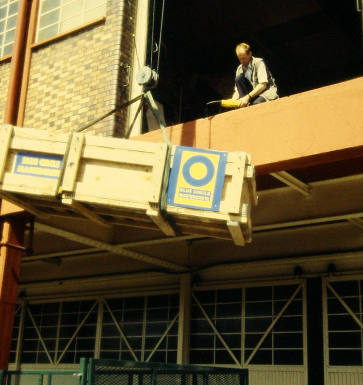 a crane lift a large crate containing the pliosaurus fossil onto the 1st floor of a building, where a man waits