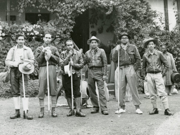 A black and white photo, taken in 1953, of a team of six South Asian men wearing hiking clothes and carrying sticks. They are posing for the photo in front of a building, possibly a hotel. One of them is Ivan Haslam. Other figures are standing and squatting in the background.