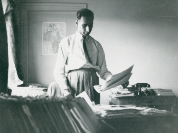 A black and white photo showing Ivan Haslam standing at a desk looking through paperwork. A map of Africa and Europe is on the wall behind him.