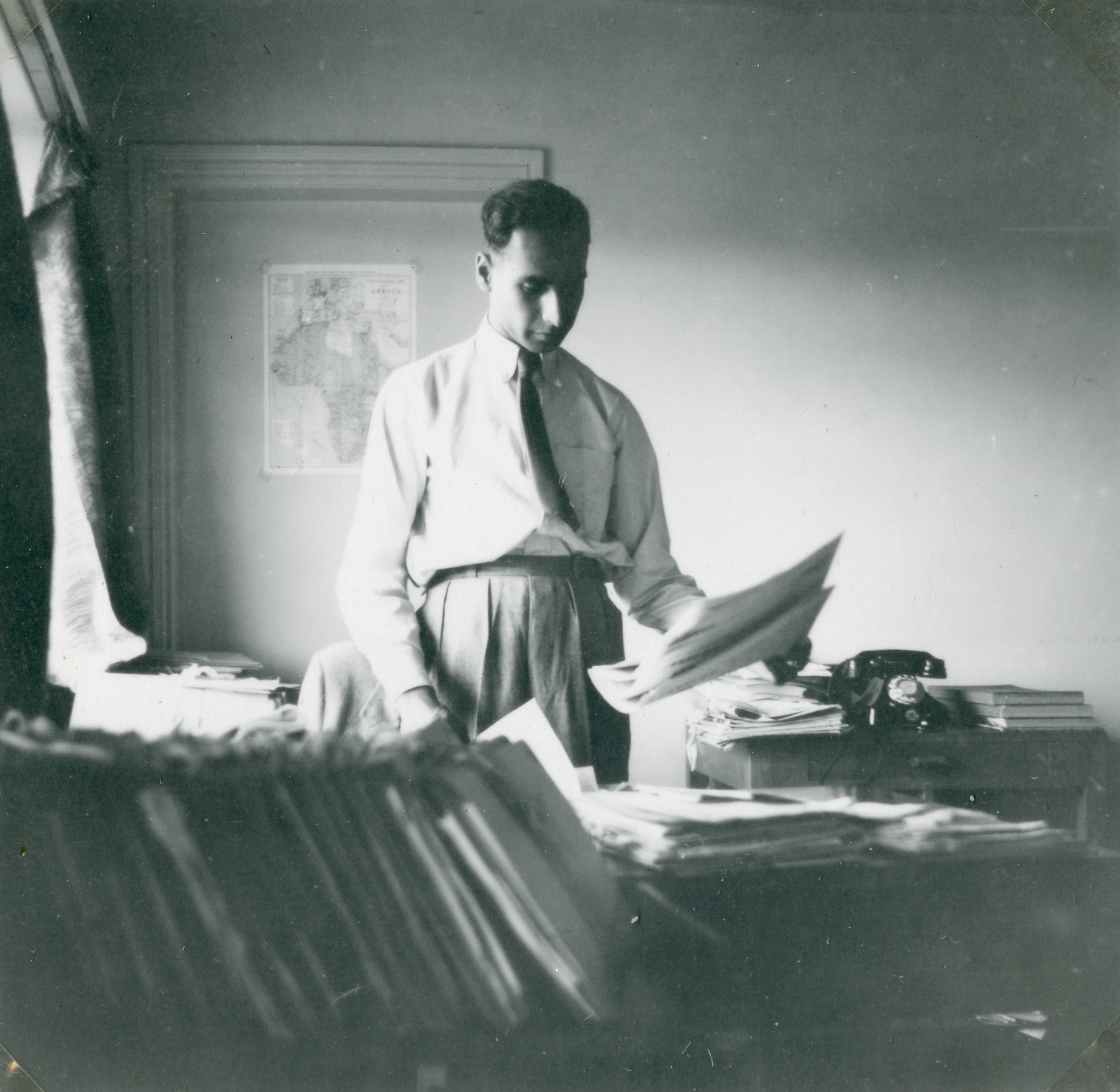 A black and white photo showing Ivan Haslam standing at a desk looking through paperwork. A map of Africa and Europe is on the wall behind him.
