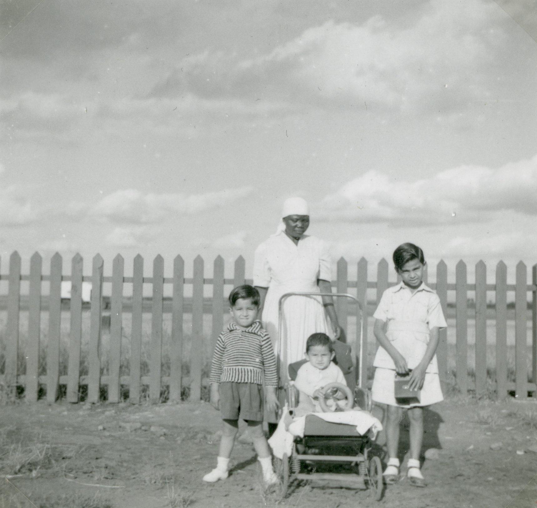 A black and white photo showing three young Asian children stand in front of a fence with an African woman in a nurse’s uniform, possibly their nanny.