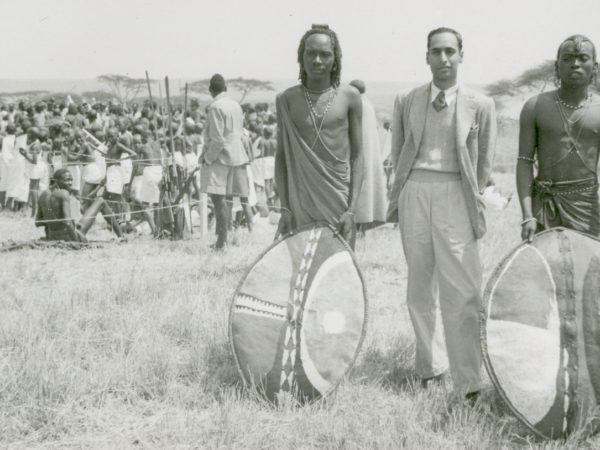 A black and white photo showing two Maasai men stand, with their shields, either side of Ivan Haslam, who is dressed in a blazer and tanktop. A crowd of other Maasai are visible in the background.