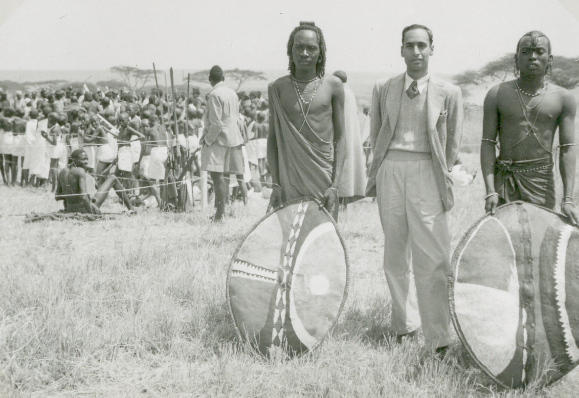 A black and white photo showing two Maasai men stand, with their shields, either side of Ivan Haslam, who is dressed in a blazer and tanktop. A crowd of other Maasai are visible in the background.