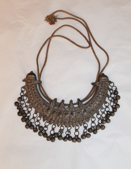 Photo of a silver necklace used by dancing women at feasts. It is made with sheet silver over a wooden form. The front is decorated with filigree work and bells are suspended from loops along the bottom. Its double cord would fasten it around a dancer's neck.