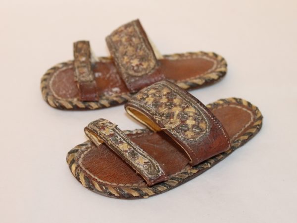 Colour photograph of a pair of child's sandals. They are made of leather. The flat sole has a decorative stitched edge. They have two straps of interlaced cream and black leather.