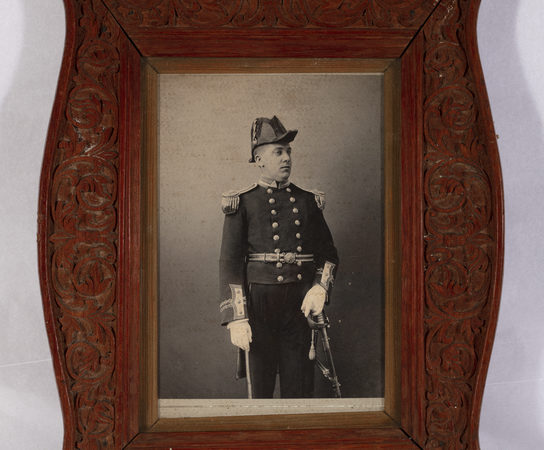Colour photograph of a black and white photograph of George Wood in his Governor's official uniform. Set in an ornate wooden frame carved in low relief with foliate scrolls. Made by prisoners in the penal colony on Ross Island in the Andaman Islands