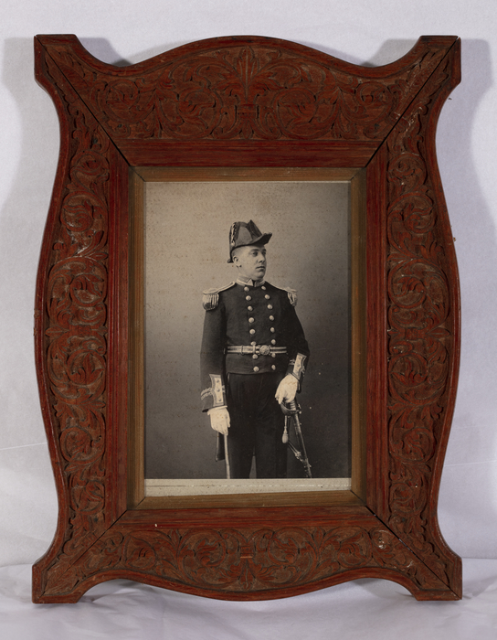 Colour photograph of a black and white photograph of George Wood in his Governor's official uniform. Set in an ornate wooden frame carved in low relief with foliate scrolls. Made by prisoners in the penal colony on Ross Island in the Andaman Islands