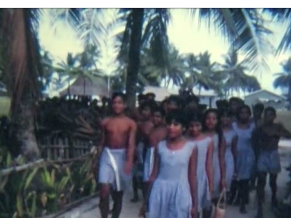 still from Mowat film: boys and girls in school uniform walking in a group down a sandy path. Palm trees line the path.