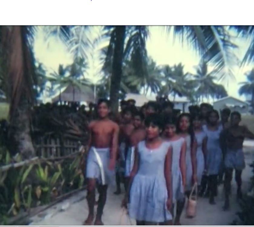 still from Mowat film: boys and girls in school uniform walking in a group down a sandy path. Palm trees line the path.