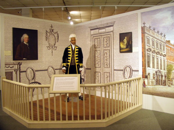 Museum display showing a Black mannequin in a white wig. Graphics imply he is in a home, acting as a servant in Georgian Britain. Bristol & Transatlantic Slavery display at Bristol Industrial Museum. (© Bristol Culture, M Shed)