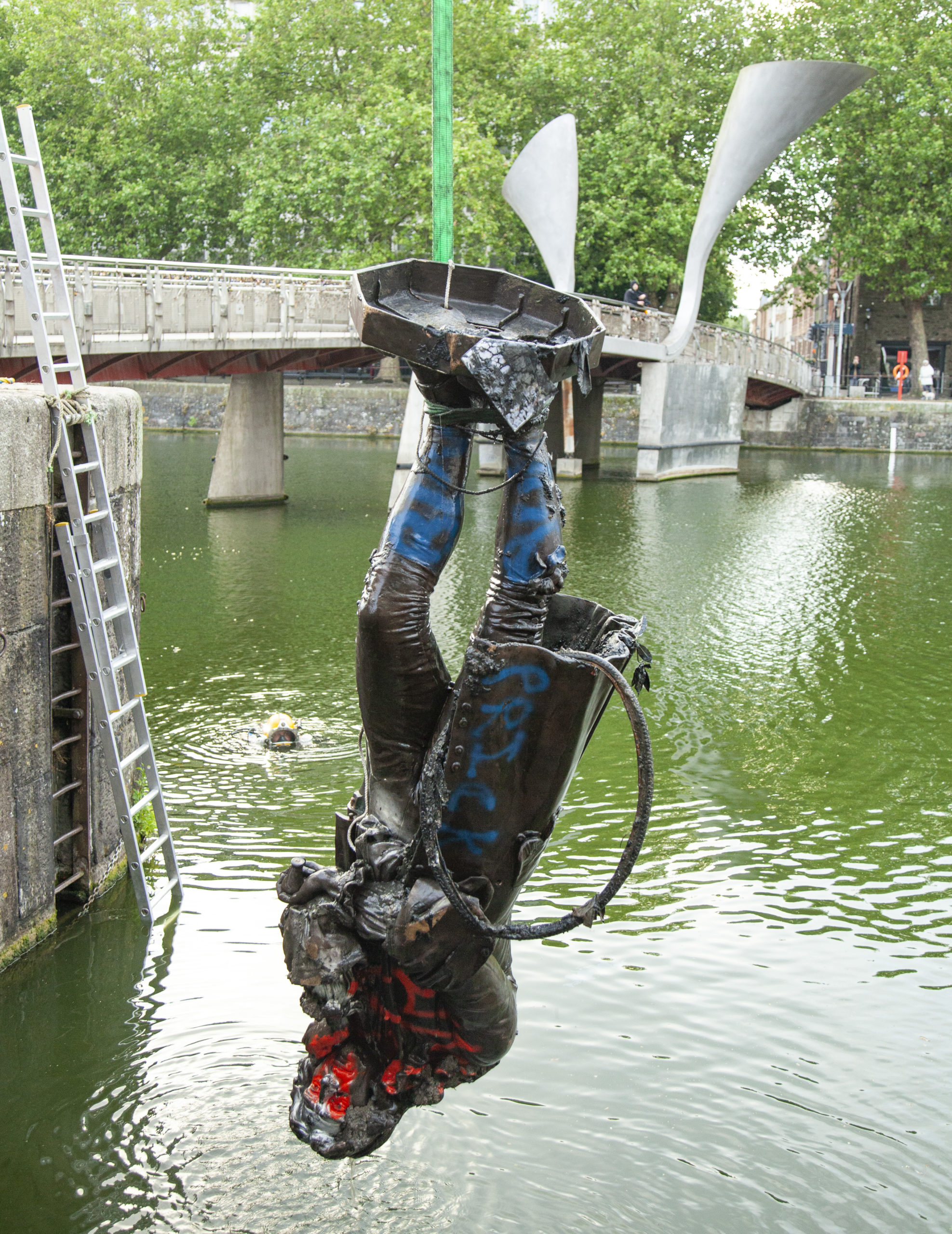 Colston statue hanging upside down, being brought up from Bristol harbour. It is painted in blue and red paint and has a tyre attached. Bridge in the background. Image Caption: Colston statue being brought up from Bristol harbour (© Bristol City Council)