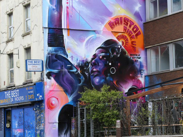 Photo of large scale graffiti on the side of a building. Painting is of a woman with a raised right fist. She wears a beret. 'BRISTOL RISE UP STAND TALL' is written above her head. Painted in purples, blues and pinks. Empty cycle racks are in the foreground.
