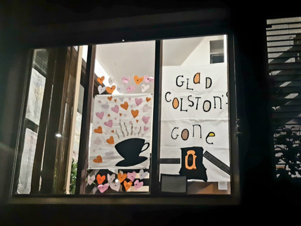 'Glad Colston's Gone' and a paper cup of tea with lots of hearts is displayed in a window. It is dark outside and the building light is on.