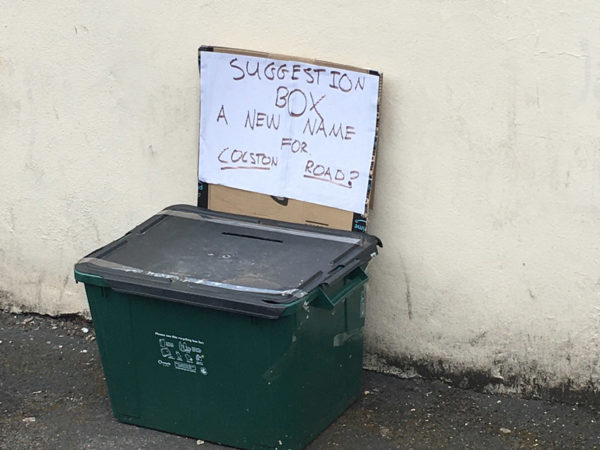 Black recycling box on a pavement. A cardboard sign on the box says ‘Suggestion box- a new name for Colston Road?’ Above it, the street name has been taped over with blue tape.