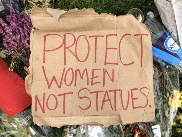 Cardboard protest placards on grass, with flower tributes. Main placard reads ‘Protect women not statues’.