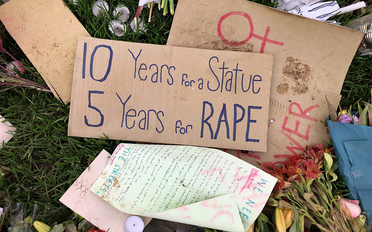 Cardboard protest placards on the ground, with candles and flowers. Main placard reads ‘10 years for a statue, 5 years for a rape’.