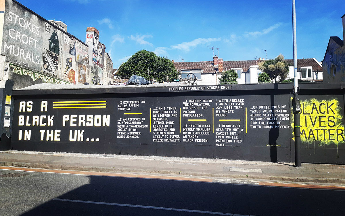 Large painted wall on a sunny day. The wall is painted with a black background with white writing, and yellow lines. Writing states ‘As a black person in the UK….’ ‘Black Lives Matter’ is painted at the other end.
