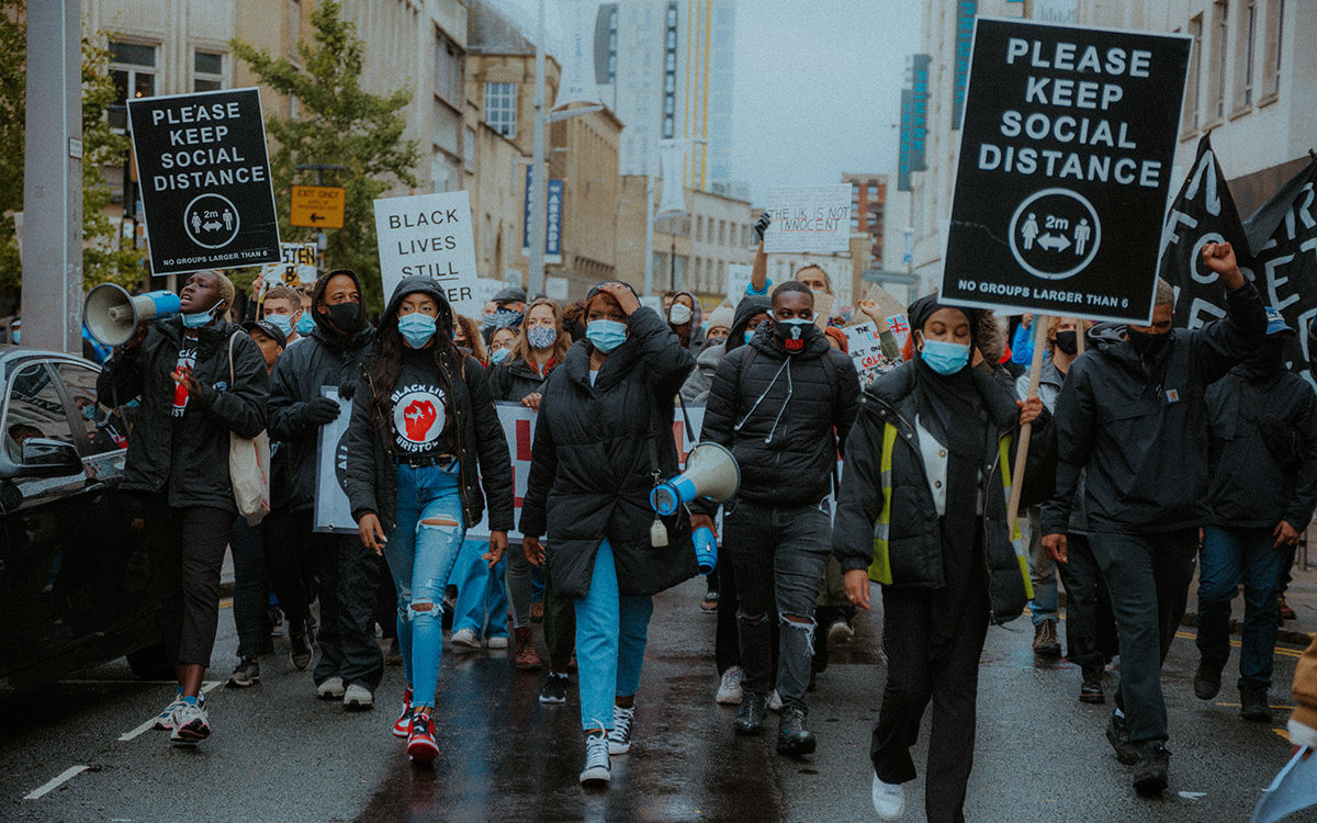 Protest march in central Bristol. Placards read ‘PLEASE KEEP SOCIAL DISTANCE’ and ‘BLACK LIVES STILL MATTER’. A Black woman on the left of the photo holds a megaphone to her mouth. Most of the crowd wear medical face masks. All Black Lives Bristol protest, Oct 2020 (© Khali Ackford)