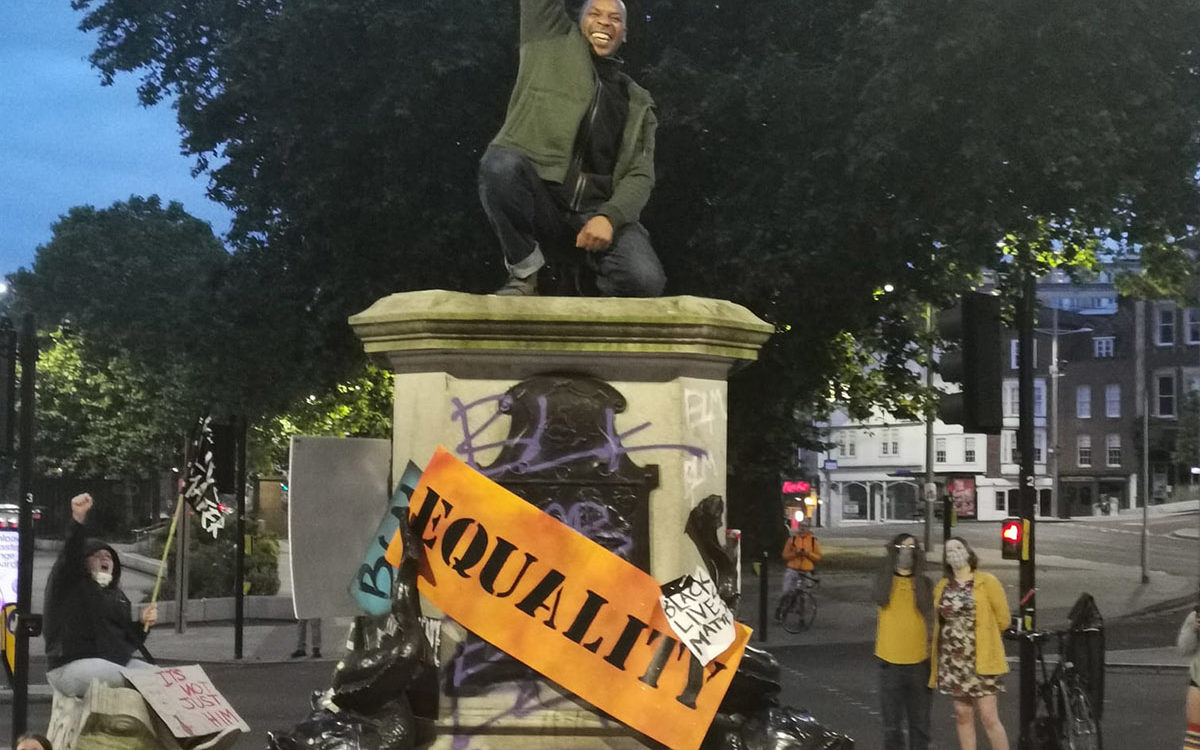 Photograph of a jubilant Black man kneeling on a statue plinth, with a raised right fist. The plinth is surrounded by placards reading ‘EQUALITY’, ‘THE UK IS NOT INNOCENT’ and ‘BLACK LIVES MATTER’.
