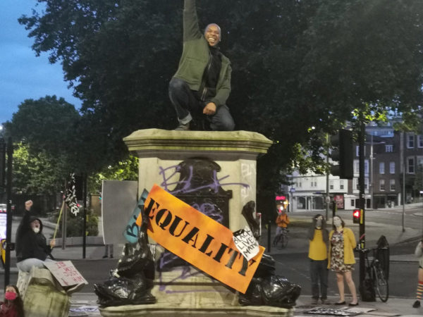 Photograph of a jubilant Black man kneeling on a statue plinth, with a raised right fist. The plinth is surrounded by placards reading ‘EQUALITY’, ‘THE UK IS NOT INNOCENT’ and ‘BLACK LIVES MATTER’.