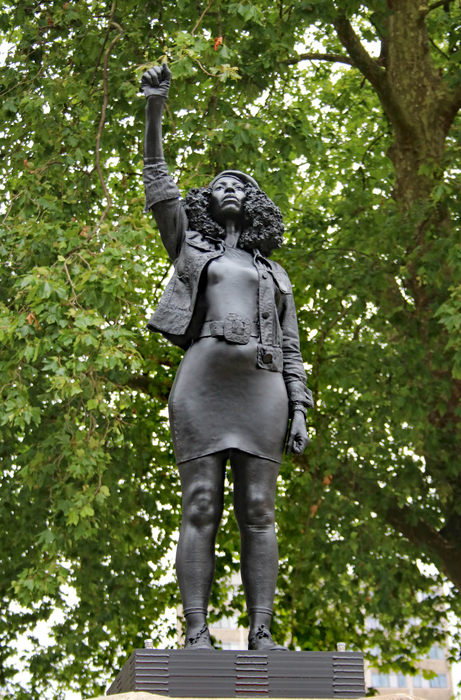Black resin statue of a woman with her right hand held in a fist above her head. Trees in the background.