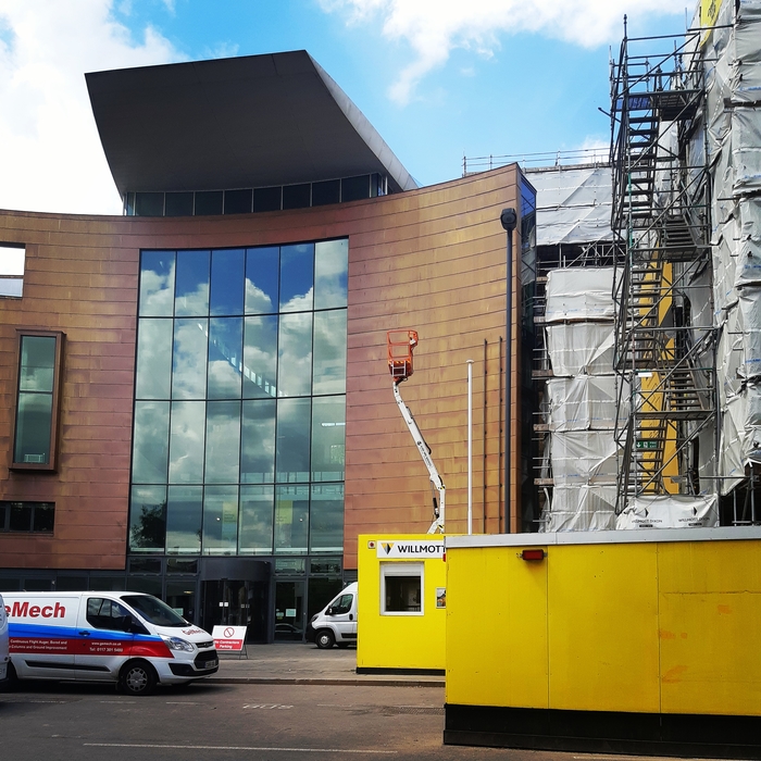 Building with scaffolding to the right, and a van parked outside. There are bright yellow building hoardings and a cherry picker. The building’s sign has been removed.